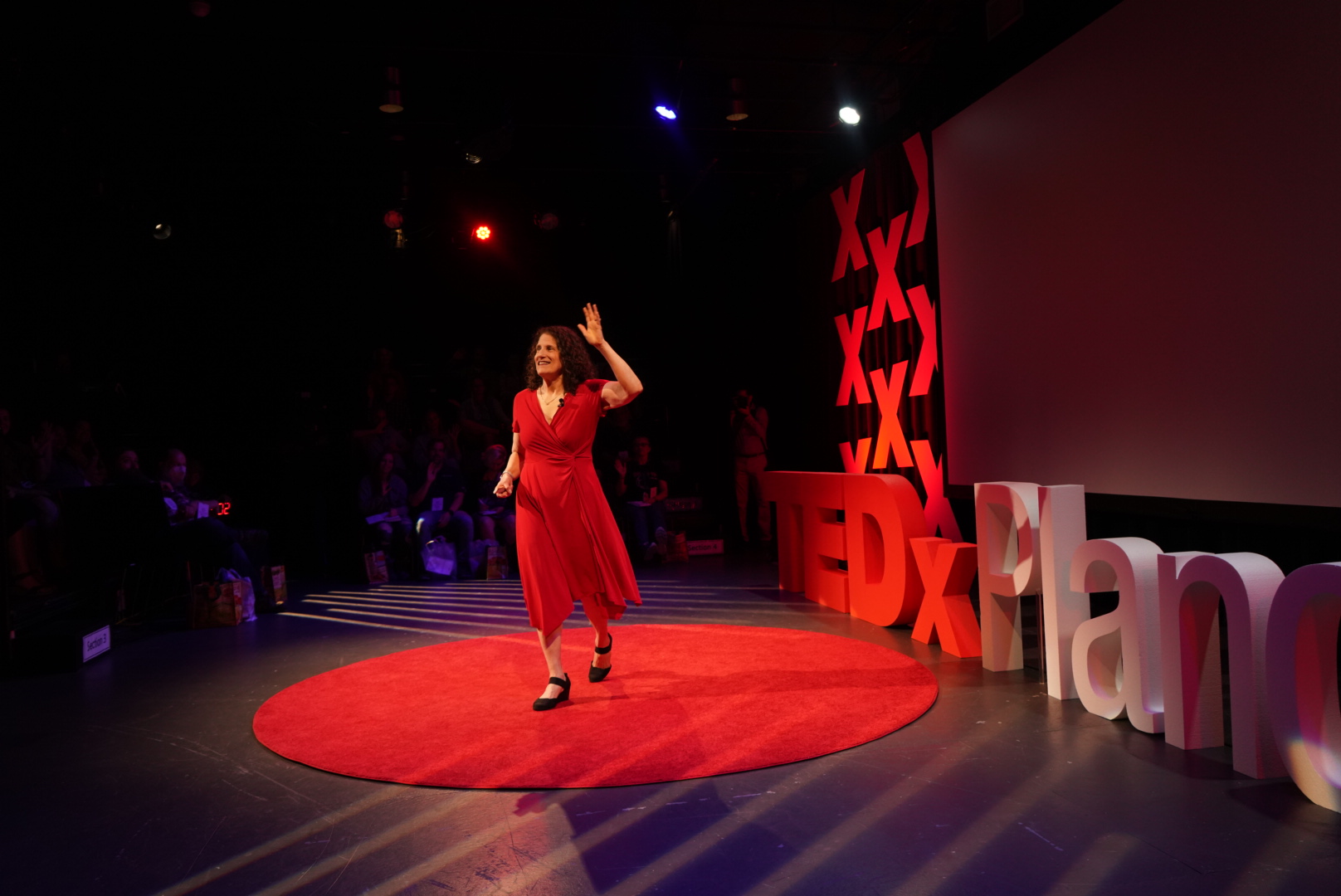 Meryl Evans waves to a full audience as she walks onstage to give a TED talk. She is wearing a bright red dress that matches the red carpet on the stage.